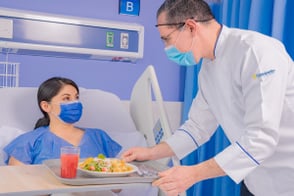 Enfermero lleva comida a mujer que reposa en cama hospitalaria